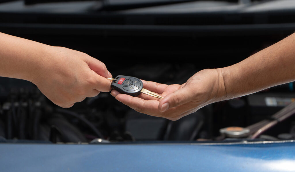 Two hands exchanging a car key in front of an open car hood.